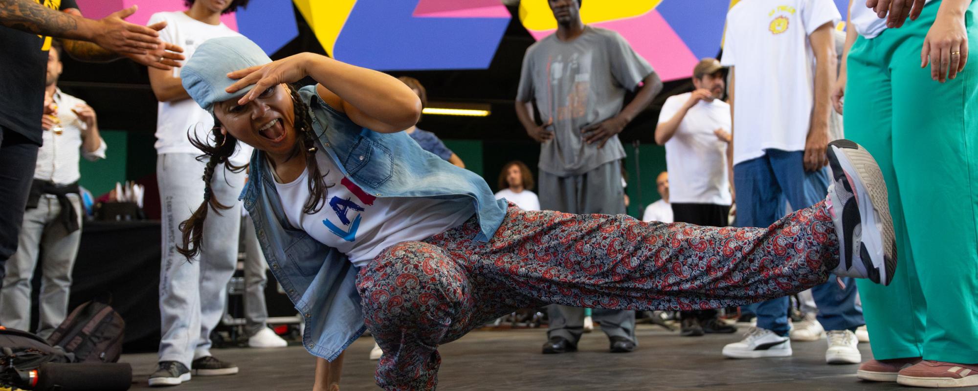 Une danseuse en plein mouvement de hip-hop. Elle a une main au sol, une jambe pliée et l'autre tendue vers le côté. Sa deuxième main couvre son front. Des personnes la soutiennent autour.