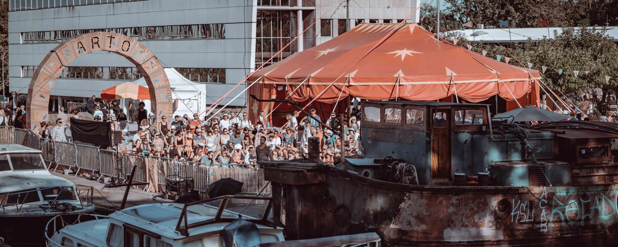 Photo d'une édition précédente du festival. Côté place du canal, les bateaux sont amarrés et le public se tient debout à côté d'un grand barnum rouge. L'entrée de l'espace est défini par une énorme arche en carton décorée de dessins avec écrit ARTO.